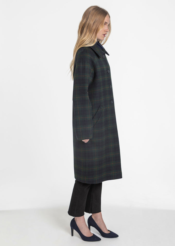 A woman modeling a long dark green and black plaid coat, shown in profile against a plain white studio background.