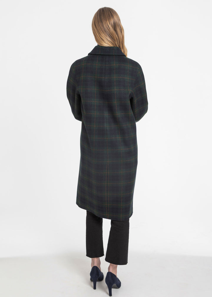 A woman shown from the back wearing a long dark green and navy plaid coat, black cropped trousers, and dark blue heels against a plain white studio background.