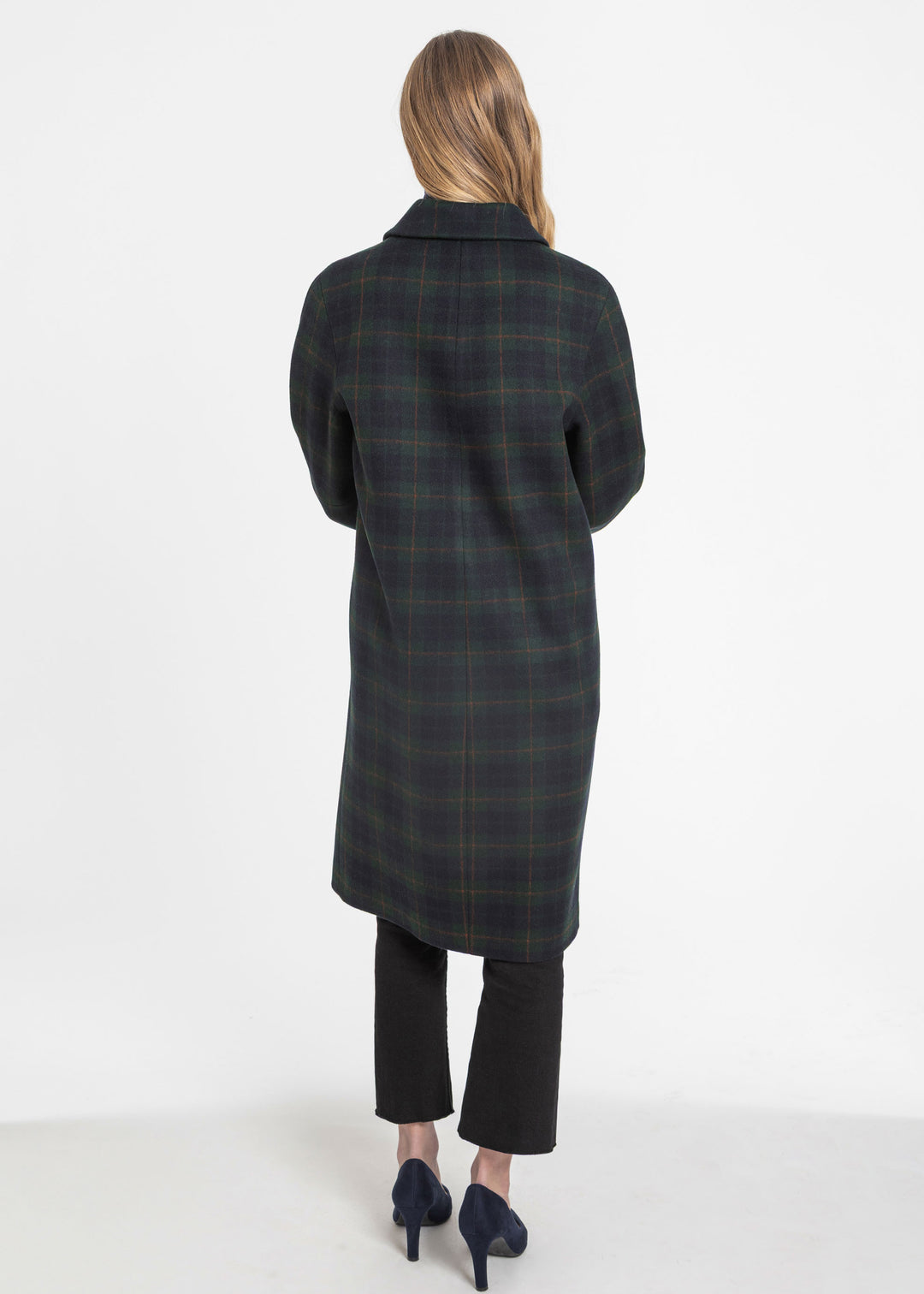 A woman shown from the back wearing a long dark green and navy plaid coat, black cropped trousers, and dark blue heels against a plain white studio background.
