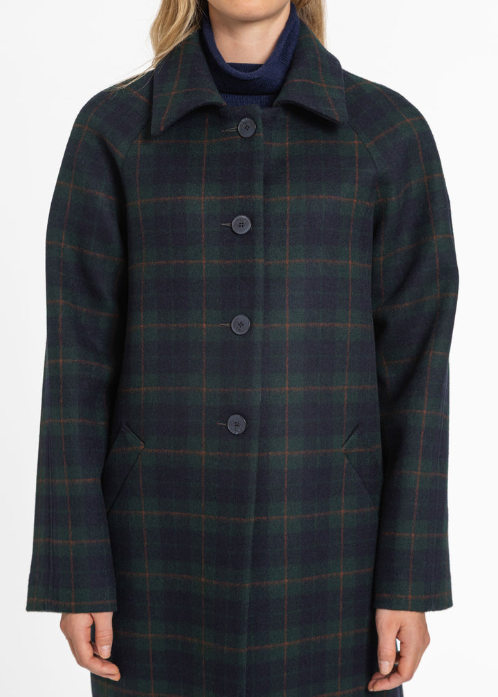 A woman wearing a dark navy and green plaid wool coat with a high collar and four front buttons, paired with a navy turtleneck underneath, shown against a plain white background.