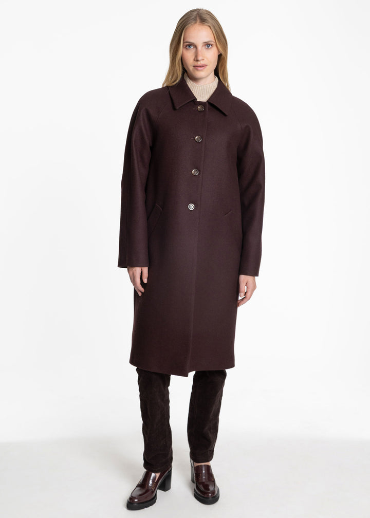 A woman modeling a long, dark mulberry wool coat with a collar and front buttons, paired with dark brown trousers and brown leather shoes, against a plain white studio background.