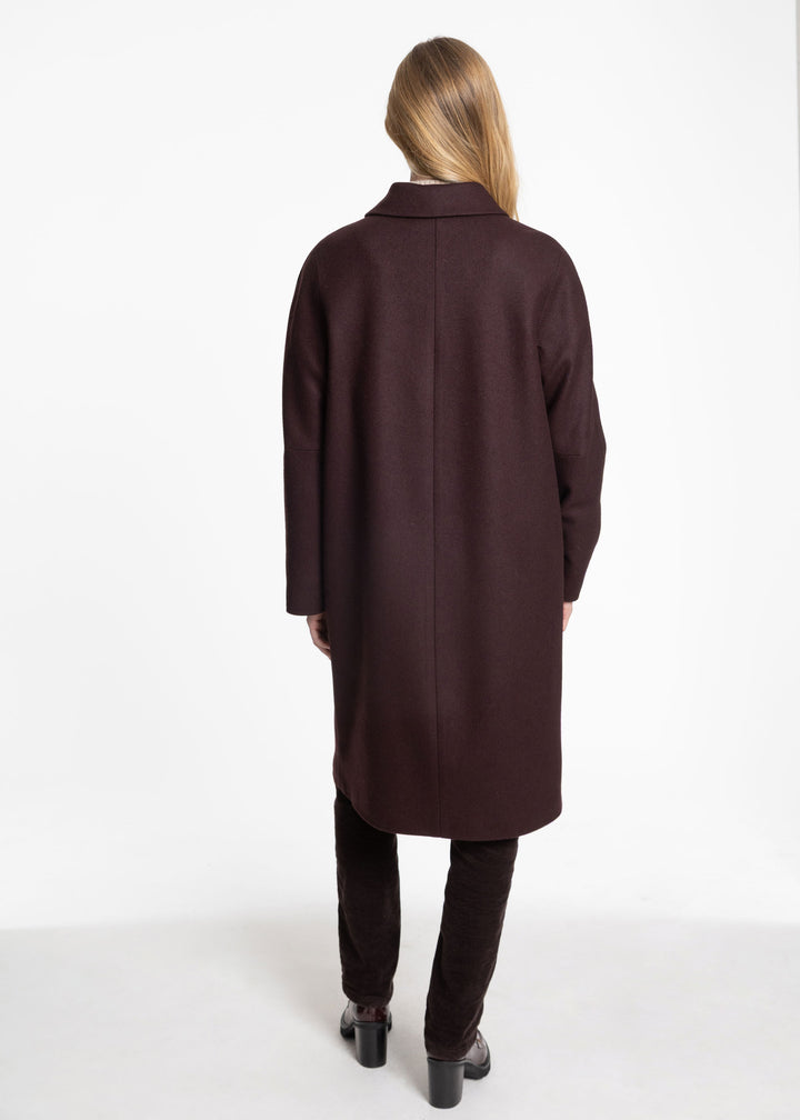 A woman viewed from behind wearing a long, dark mulberry wool coat, dark brown trousers, and black shoes against a plain white studio background.