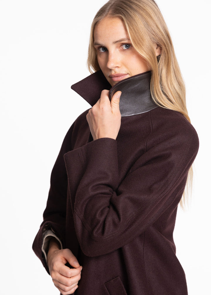 A blonde woman wearing a dark mulberry wool coat with a contrasting brown leather under collar, posing against a solid white background.