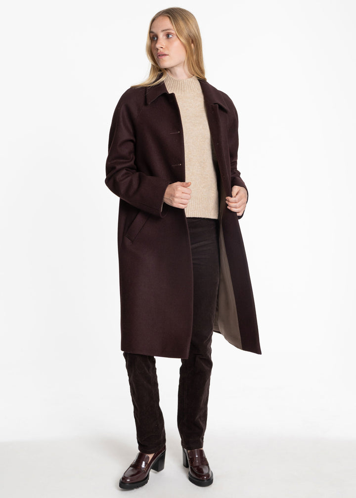 A woman modeling a long, dark mulberry coat over a beige turtleneck sweater, black pants, and dark brown leather shoes, photographed against a plain white studio background.