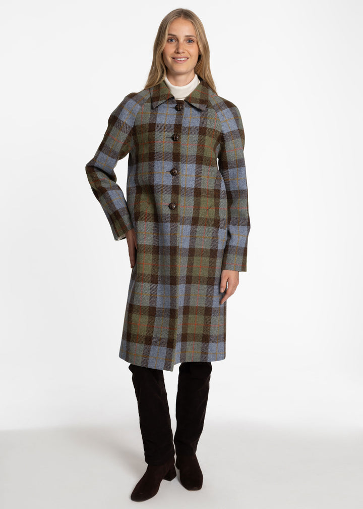 A woman modeling a knee-length plaid coat with a collar and front buttons, worn over a white top and dark brown boots, photographed against a plain white studio background.