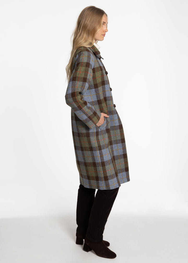 A woman modeling a knee-length plaid coat in side profile against a plain white studio background. The coat features a muted plaid pattern with blue, green, brown, and beige tones, and is paired with black pants and dark brown ankle boots.