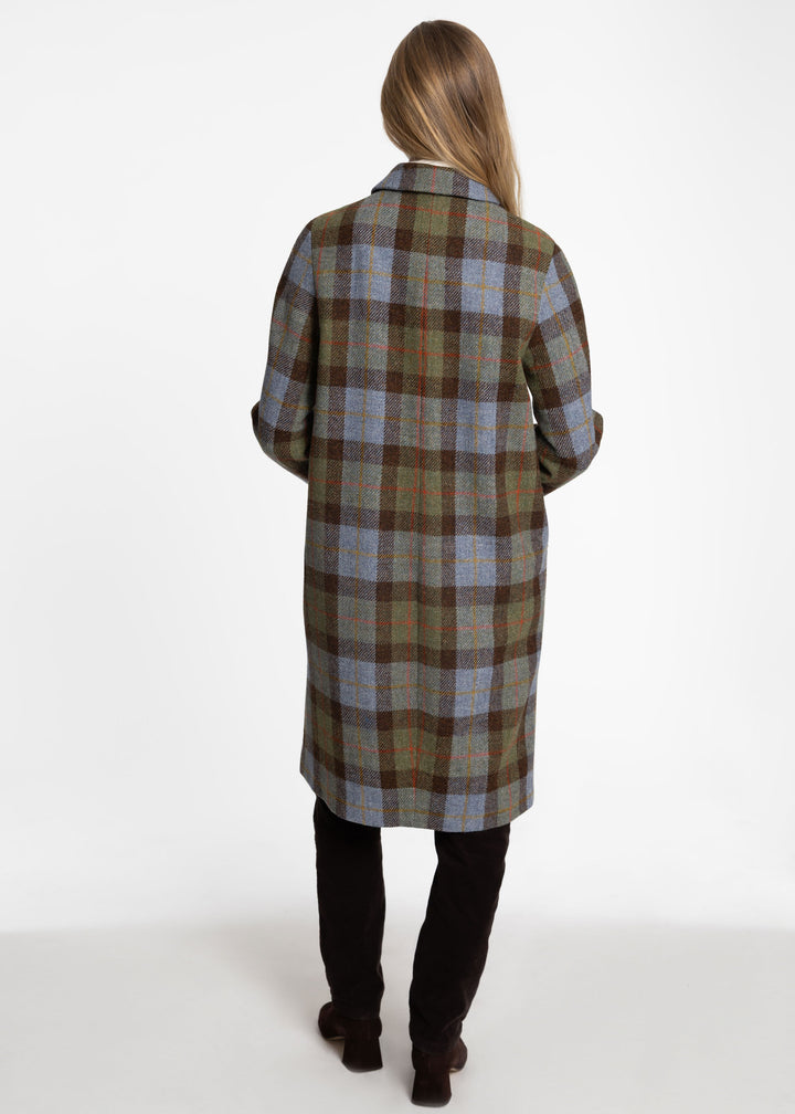 A woman viewed from behind wearing a long plaid coat in muted green, brown, and blue tones, paired with black pants and dark boots, against a plain white studio background.