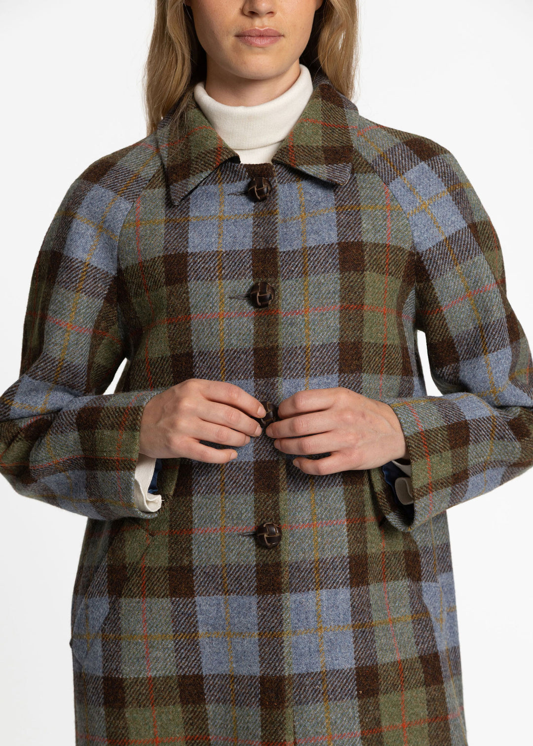 A woman wearing a long plaid coat over a white turtleneck, shown from the mouth down against a plain light background.
