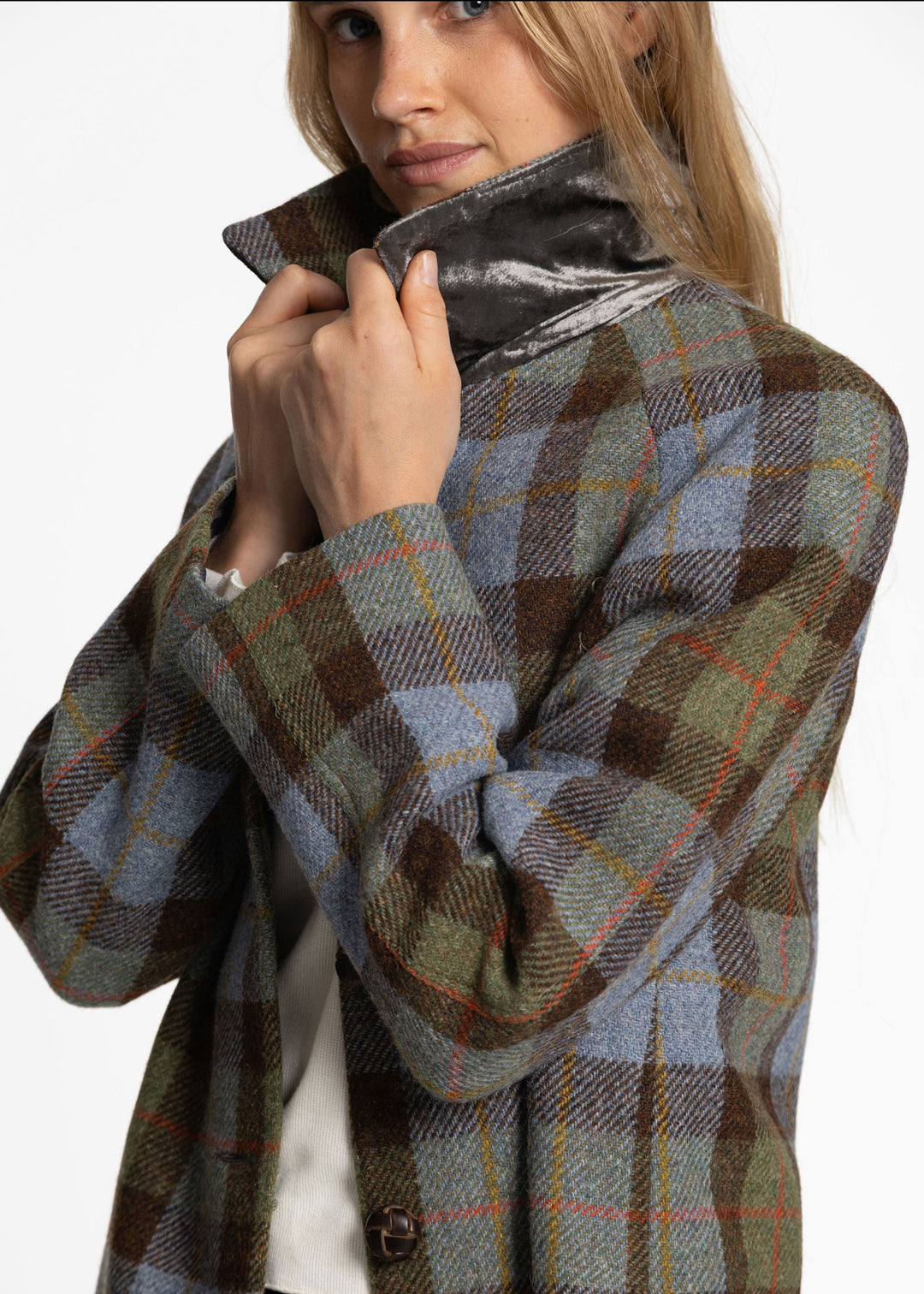 A close-up of a woman wearing a high-neck plaid coat in muted brown, blue-gray, and olive tones, with a shiny dark collar, against a plain light background.