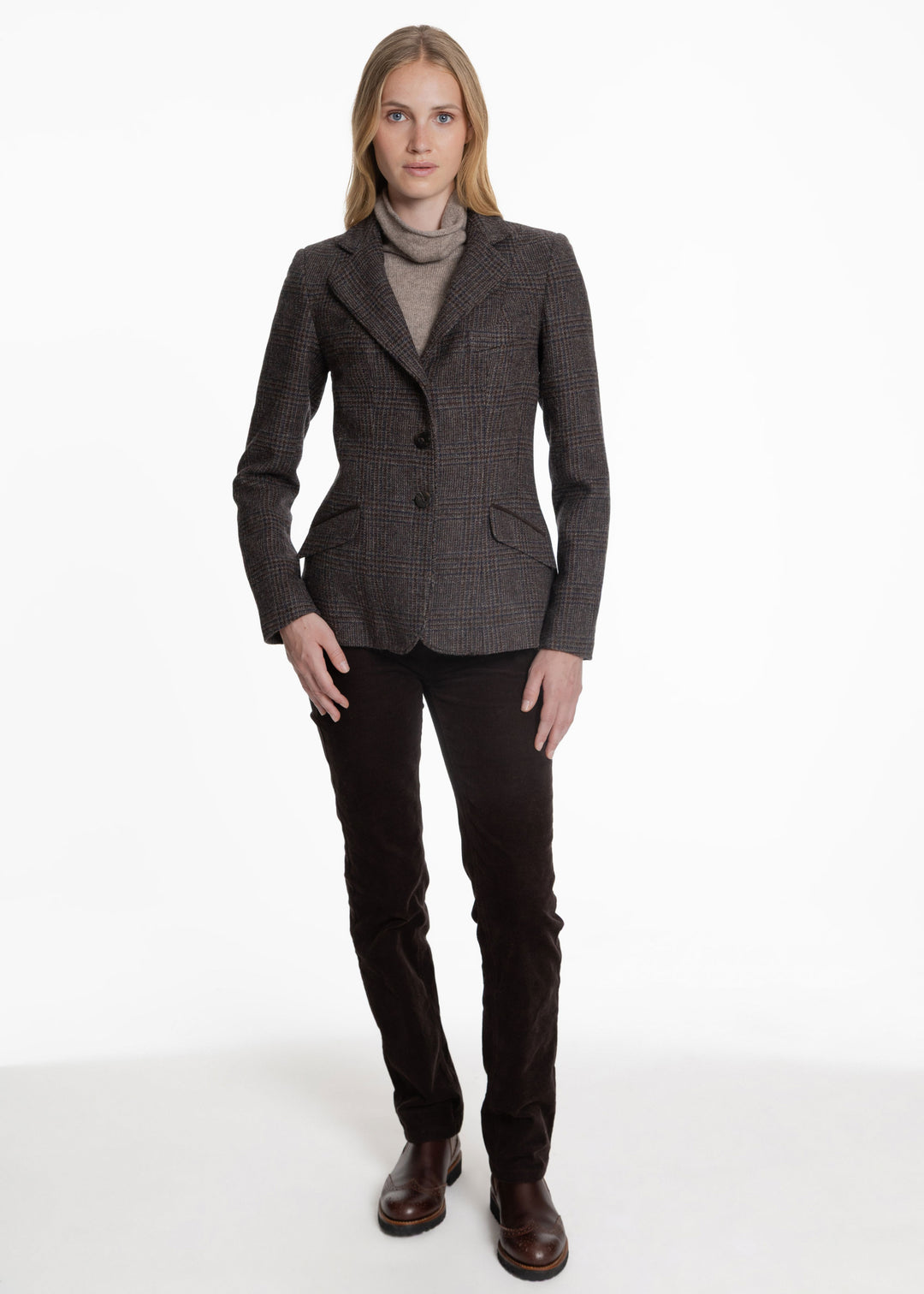 A woman modeling a dark brown and gray check jacket over a gray turtleneck sweater, paired with dark brown trousers and brown leather boots, against a plain white studio background.