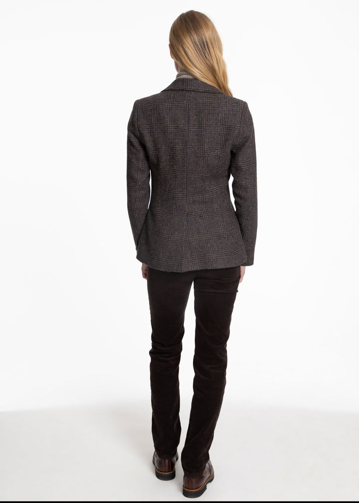 A woman viewed from the back wearing a dark brown and black check jacket, dark brown trousers, and brown leather shoes, standing against a plain white studio background.