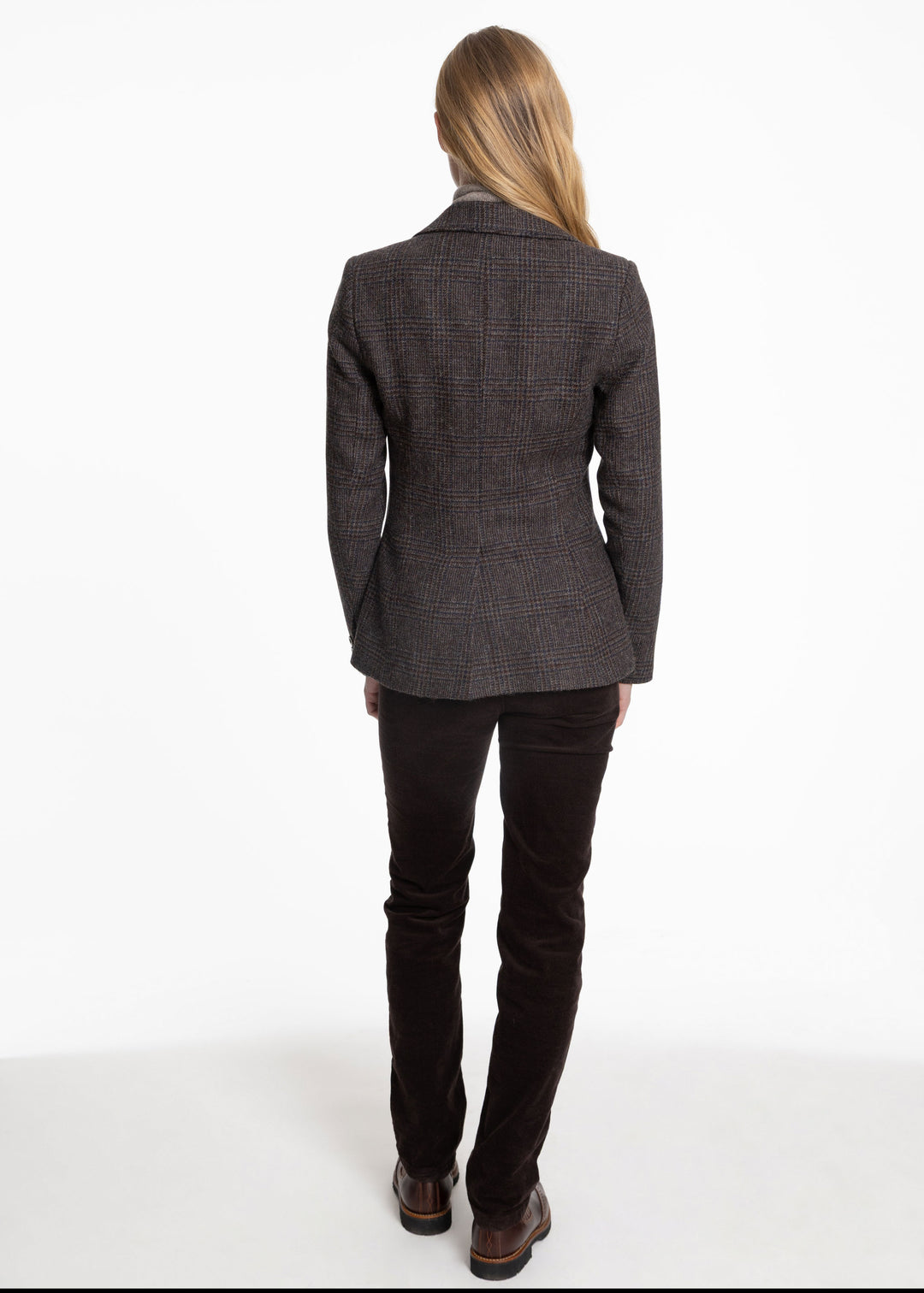 A woman viewed from the back wearing a dark brown and black check jacket, dark brown trousers, and brown leather shoes, standing against a plain white studio background.