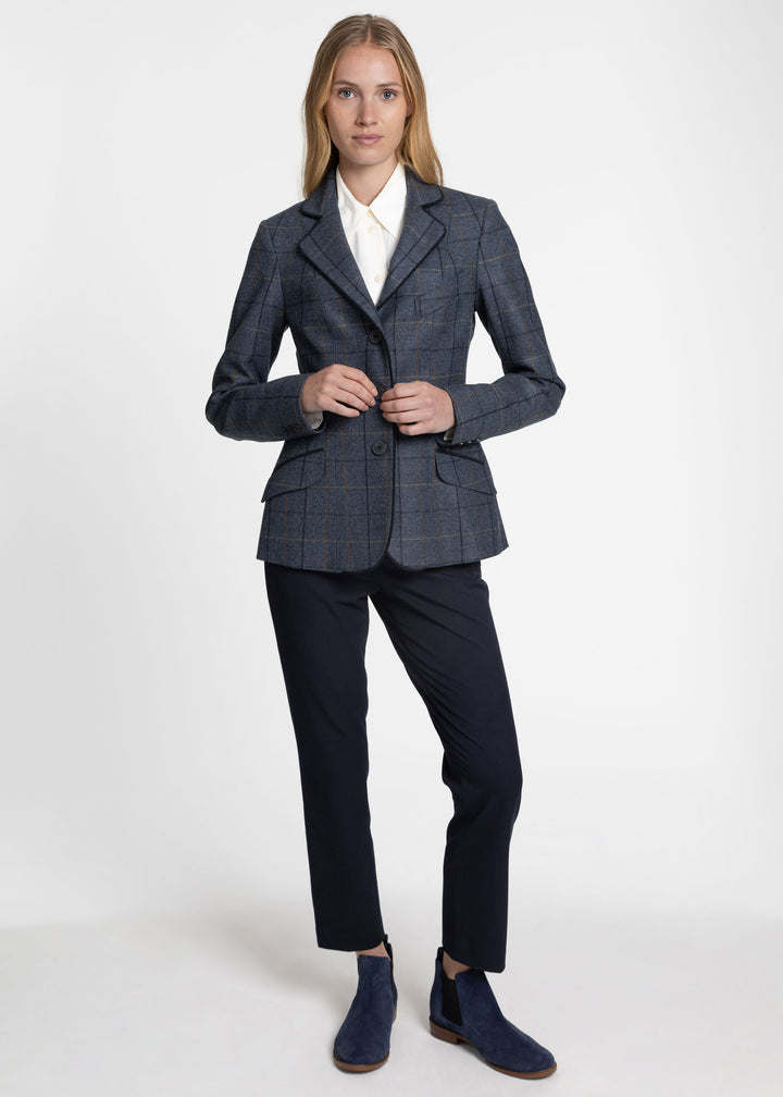A woman modeling a tailored jacket in a dark blue plaid pattern, paired with dark navy trousers and navy blue suede boots, against a plain light gray studio background.