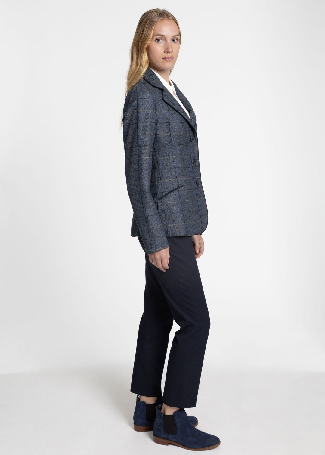 A woman modeling a tailored navy blue plaid jacket with dark navy trousers and navy Chelsea-style boots, standing against a plain light gray studio background.