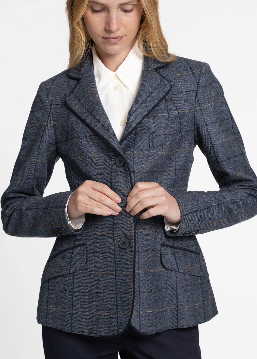 A woman wearing a tailored windowpane plaid jacket in dark blue with thin orange lines, styled over a white collared shirt. She is shown from the head down to the waist, adjusting the front buttons of the jacket against a clean white background.