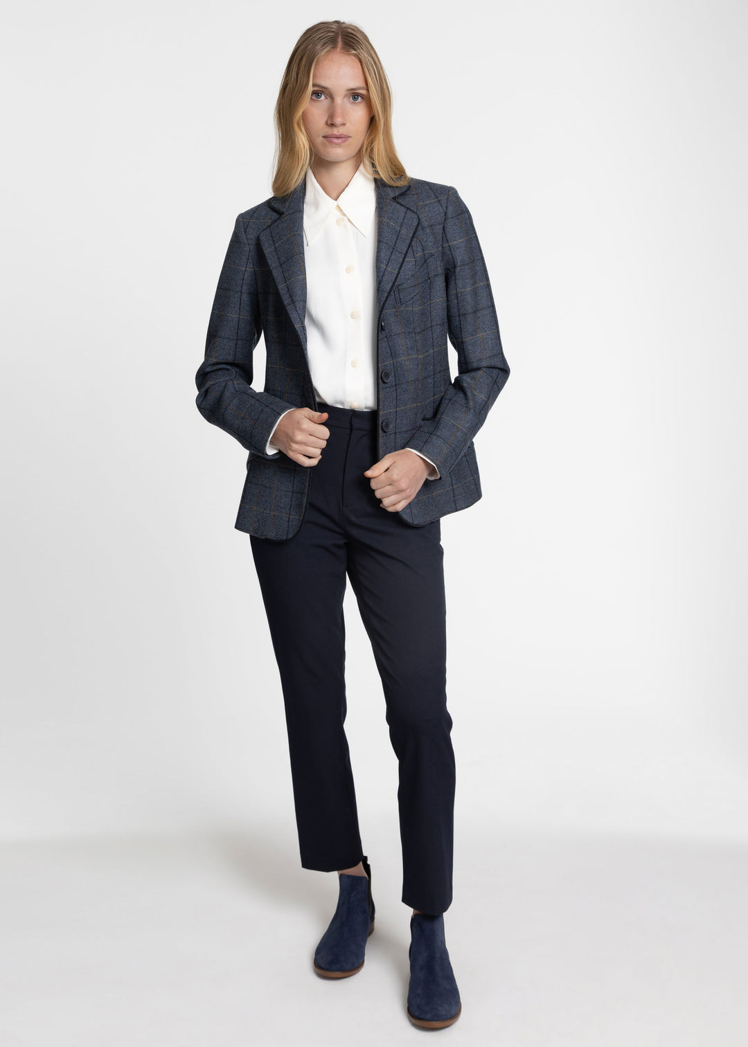 A woman modeling a tailored navy blue plaid jacket with dark navy trousers, a white collared shirt, and navy ankle boots against a light gray studio background.