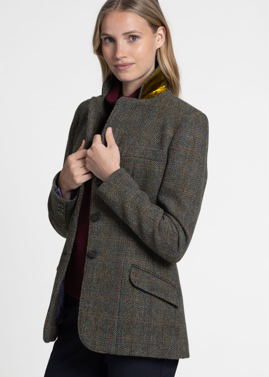 A woman wearing a green tweed jacket with a subtle plaid pattern, styled over a burgundy top and a gold collar insert, against a plain light gray studio background.