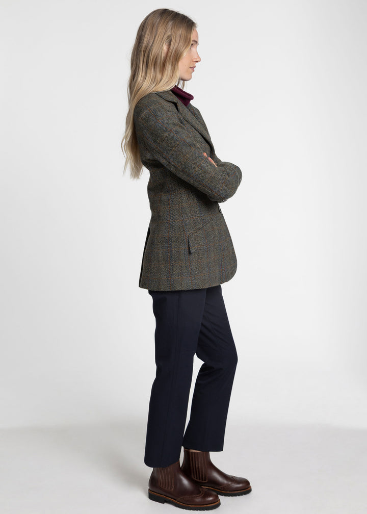 A woman stands in profile wearing a green herringbone jacket, dark navy trousers, and brown leather ankle boots against a plain light gray studio background.