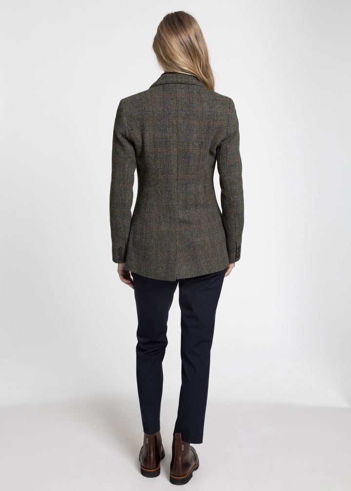 A woman viewed from the back wearing a tailored green tweed jacket, paired with dark navy trousers and brown leather ankle boots, against a plain white studio background.