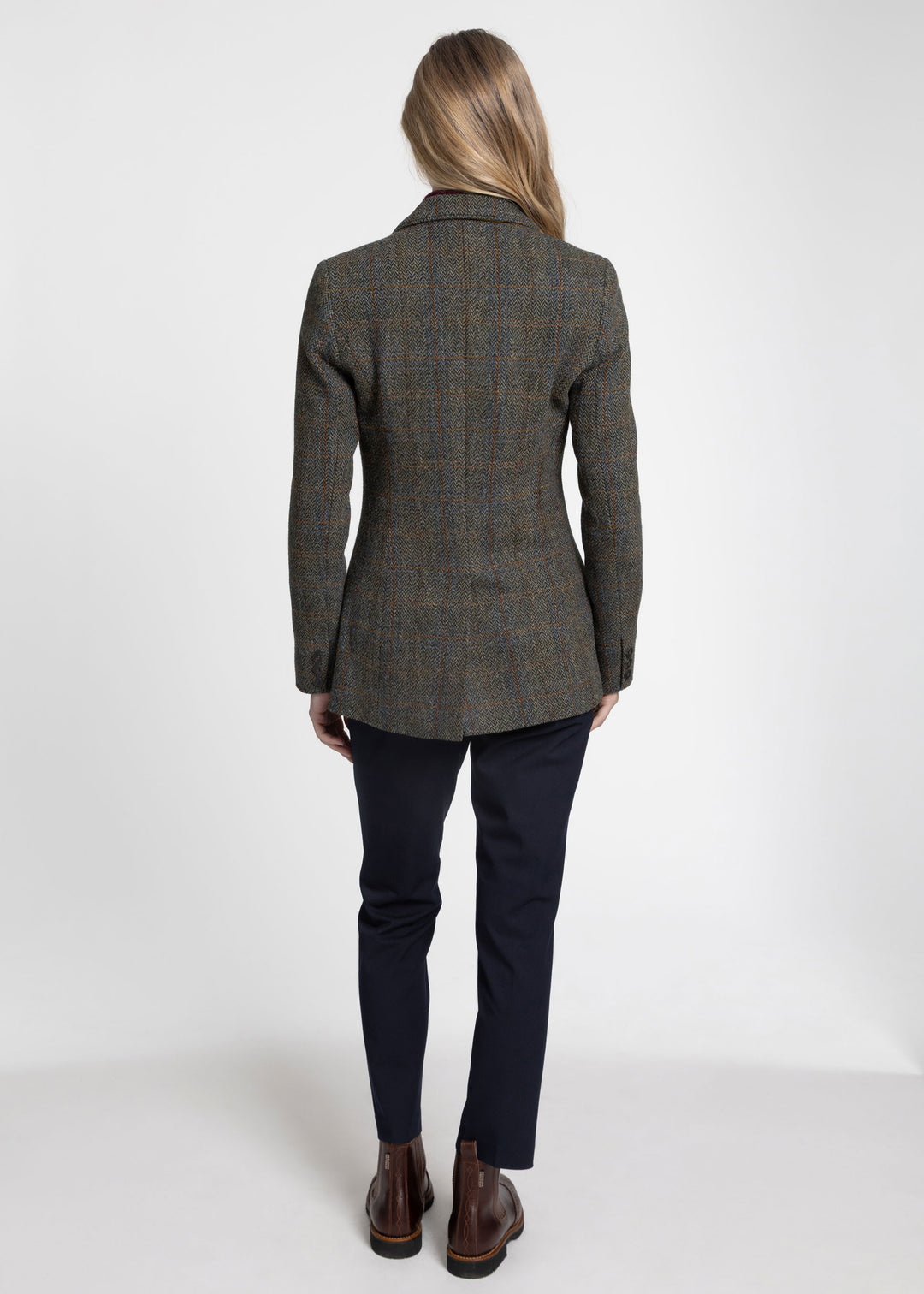 A woman viewed from the back wearing a tailored green tweed jacket, paired with dark navy trousers and brown leather ankle boots, against a plain white studio background.