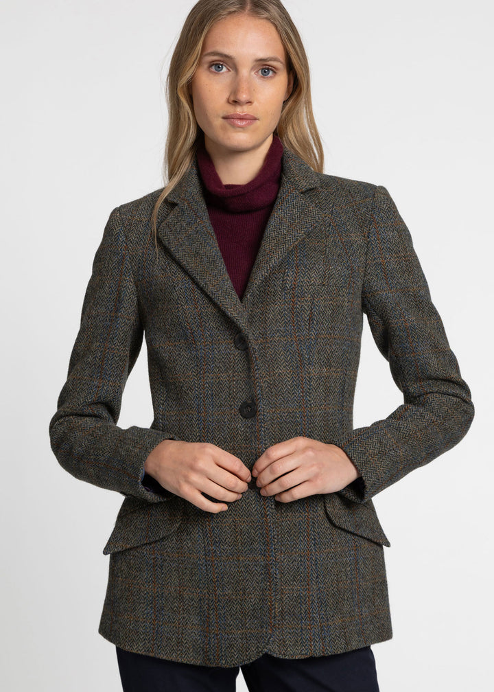 A woman wearing a tailored tweed blazer in a dark green herringbone pattern over a burgundy turtleneck, photographed against a plain light gray studio background.