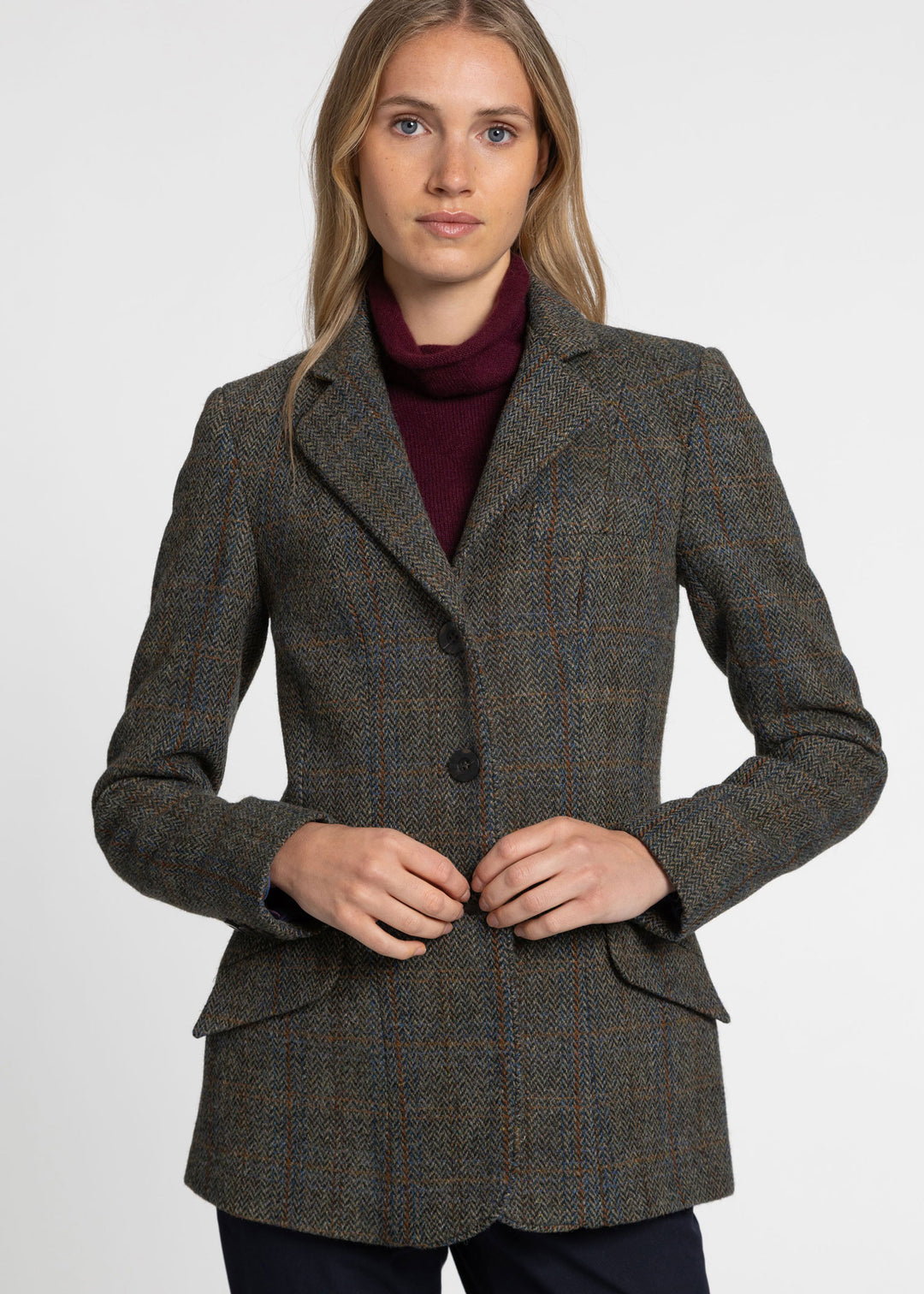 A woman wearing a tailored tweed blazer in a dark green herringbone pattern over a burgundy turtleneck, photographed against a plain light gray studio background.