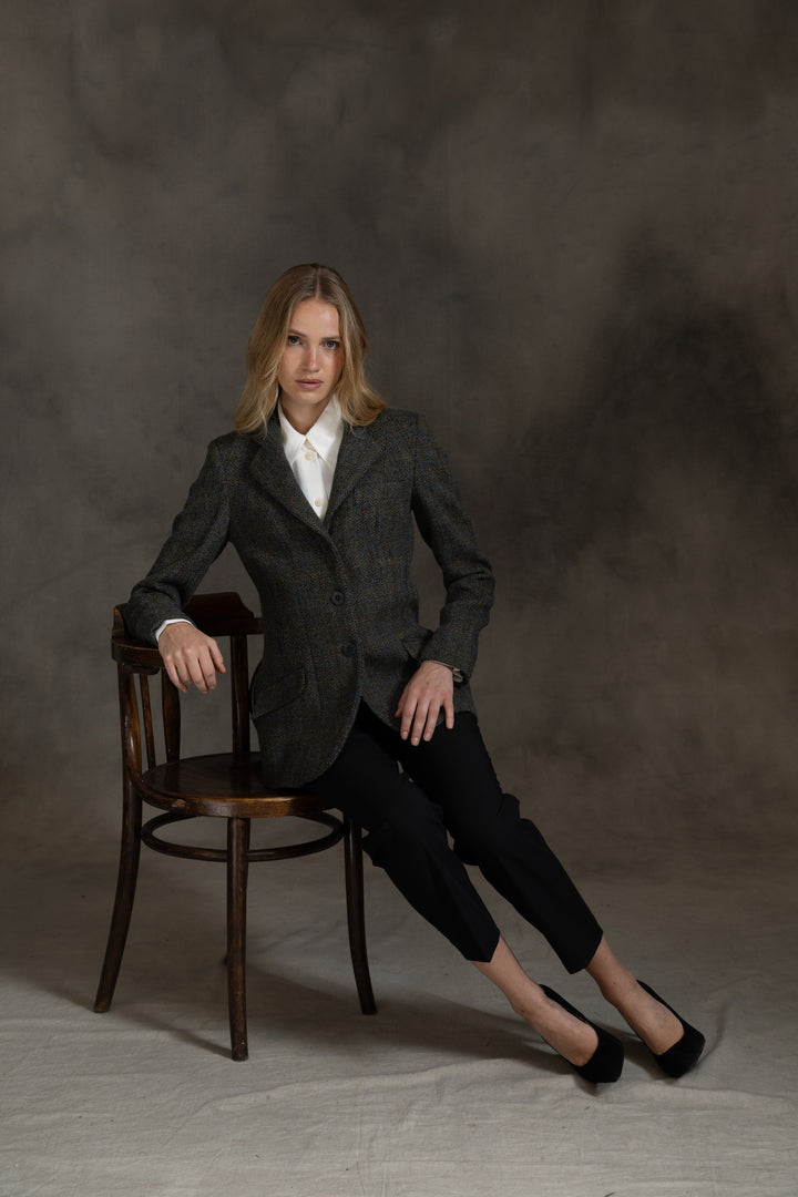 A woman sits on a dark wooden chair against a textured gray studio backdrop, wearing a dark green patterned jacket over a white collared shirt, black cropped pants, and black heels.