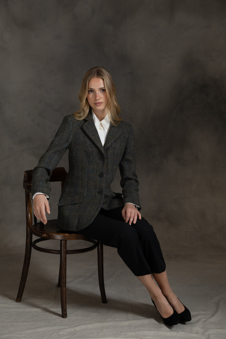A woman sits on a wooden chair wearing a dark green and black herringbone jacket over a white collared shirt, paired with black cropped pants and black heels, against a textured gray studio backdrop.