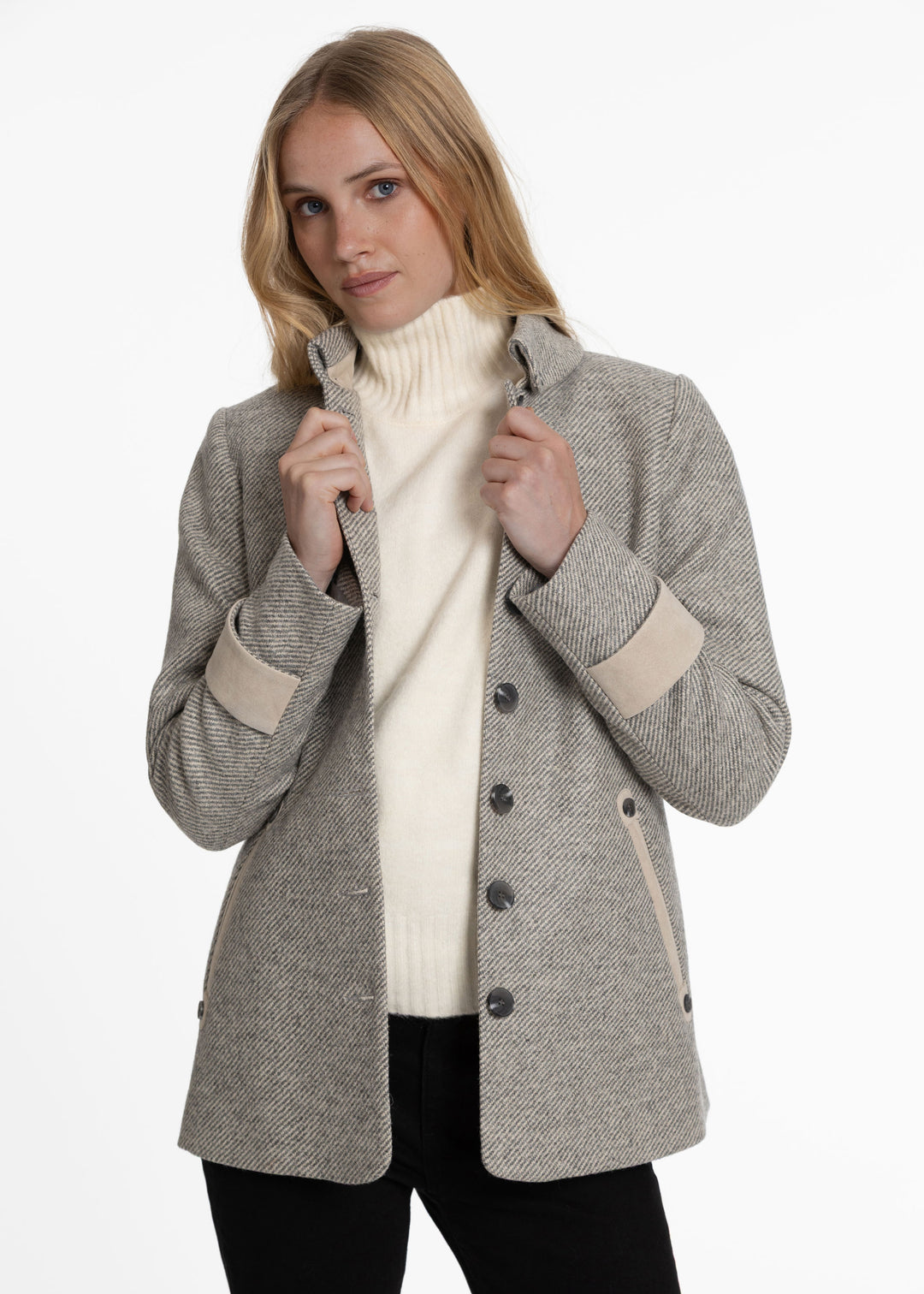 A woman wearing a light gray twill wool-blend jacket with beige contrast trim on the cuffs and pockets, layered over a cream turtleneck sweater and black pants, against a plain white studio background.