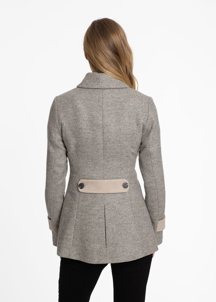 A woman viewed from the back wearing a light gray and beige twill jacket with beige pockets and a beige waist belt, paired with black pants, against a plain white background.