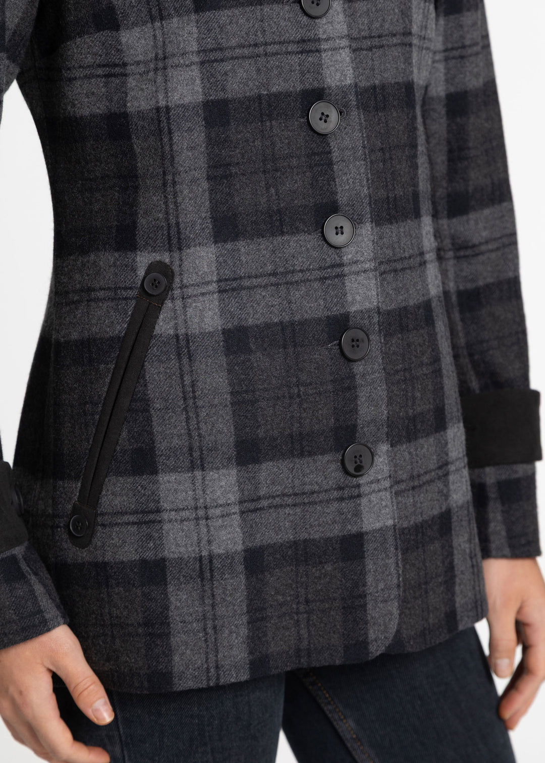 Close-up of a person wearing a dark gray and black plaid jacket with black buttons and a black pocket, paired with dark blue jeans.