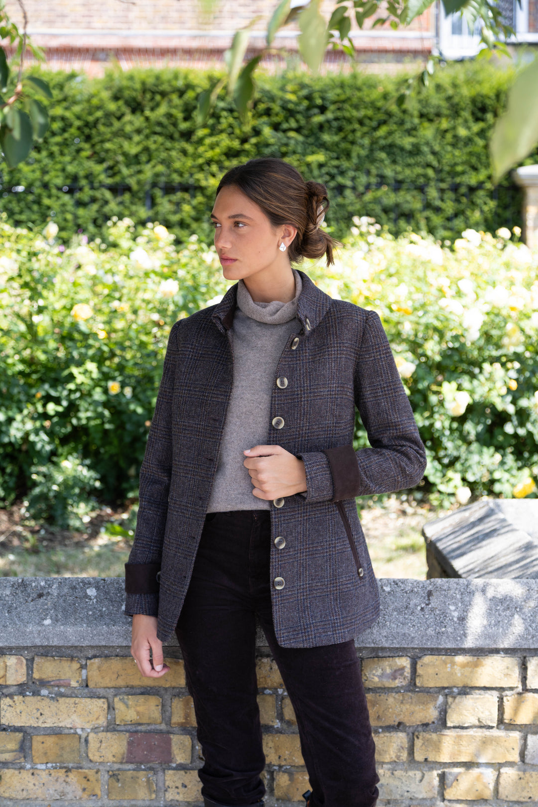A woman wearing a dark brown and black patterned tweed jacket over a gray turtleneck sweater and black trousers, standing outdoors against a brick wall with a green hedge background.