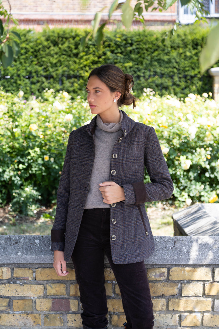 A woman wearing a dark brown and black patterned tweed jacket over a gray turtleneck sweater and black trousers, standing outdoors against a brick wall with a green hedge background.