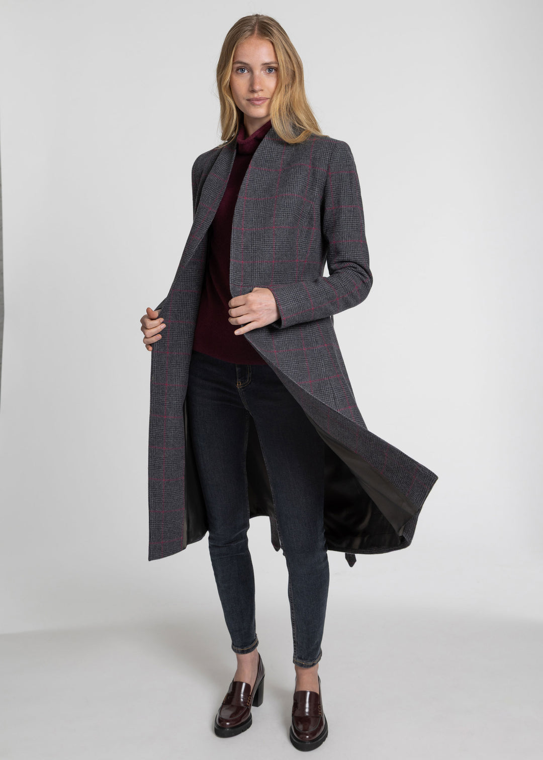 A woman modeling a long charcoal gray windowpane plaid coat over a burgundy turtleneck, dark skinny jeans, and brown leather loafers, photographed against a light gray studio background.