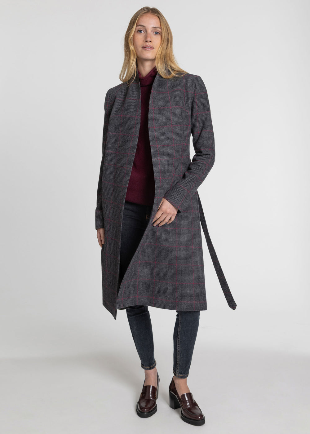 A woman modeling a long gray windowpane plaid coat with a dark belt, worn over a burgundy top and dark jeans, on a light gray studio background.