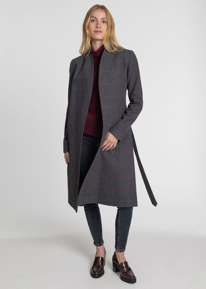 A woman modeling a long gray windowpane plaid coat with a dark belt, worn over a burgundy top and dark jeans, on a light gray studio background.