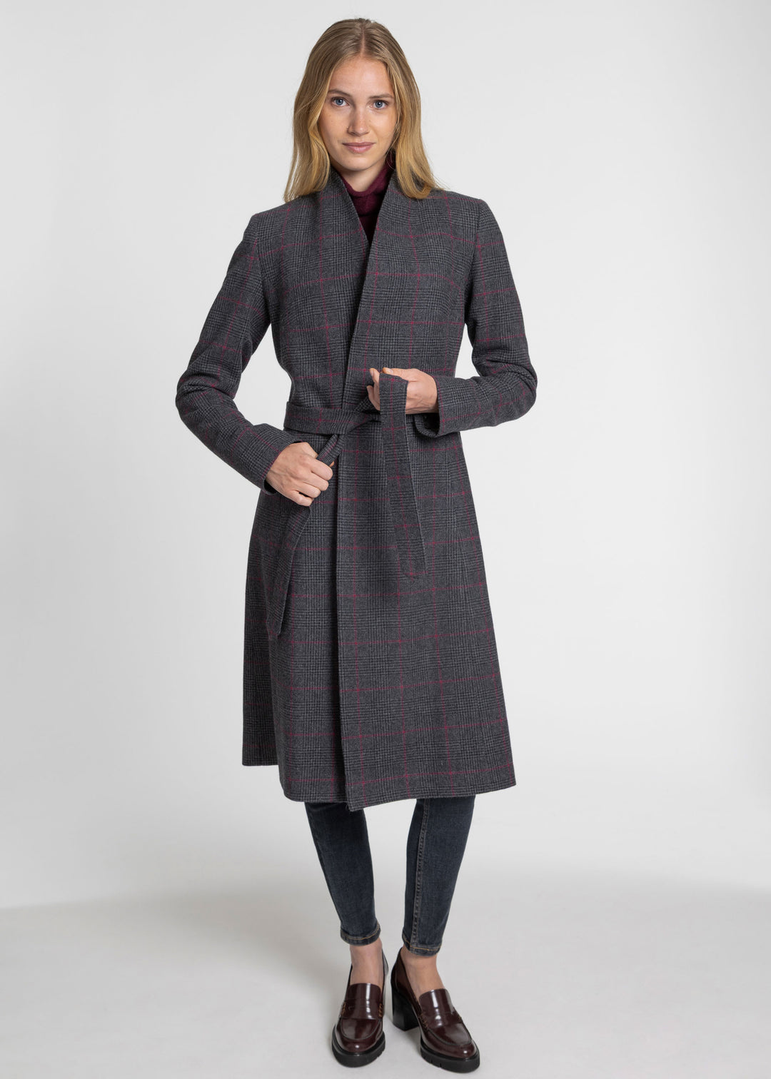 A woman modeling a dark gray belted windowpane coat with a subtle red check pattern, paired with dark gray jeans and brown leather loafers, against a light gray studio background.