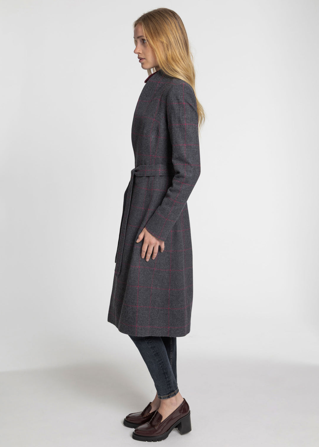 A woman modeling a long dark gray windowpane plaid coat with a matching waist tie, paired with dark jeans and brown loafers, shown from a side profile against a light gray studio background.