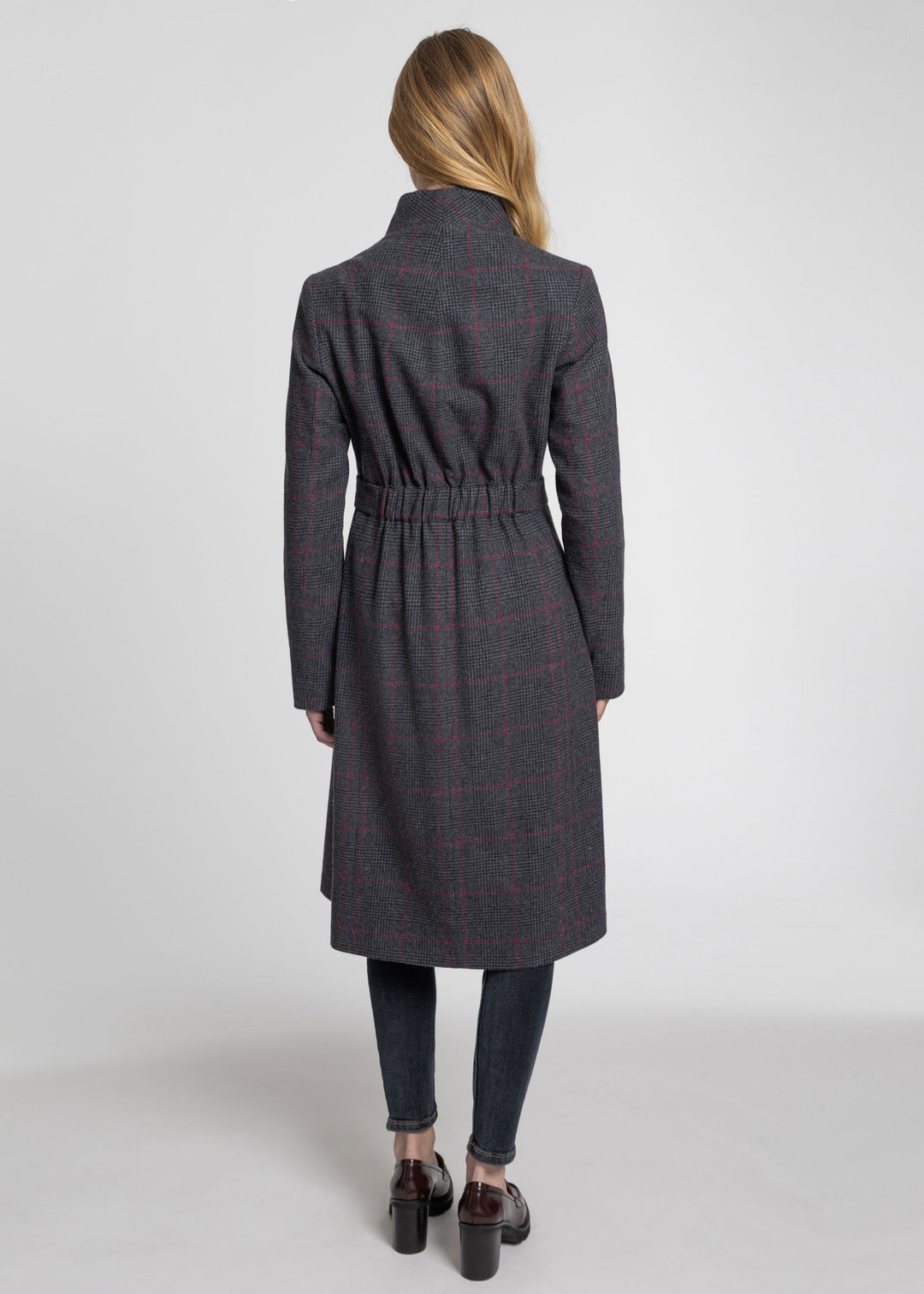 A woman viewed from the back wearing a dark gray belted coat with a subtle red windowpane plaid pattern, paired with dark jeans and brown shoes, against a light gray studio background.