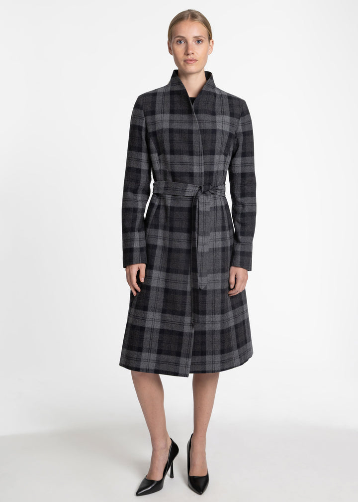 A woman modeling a long-sleeved, knee-length plaid coat with a shawl collar and matching waist tie, paired with black pointed-toe heels, against a plain white studio background.