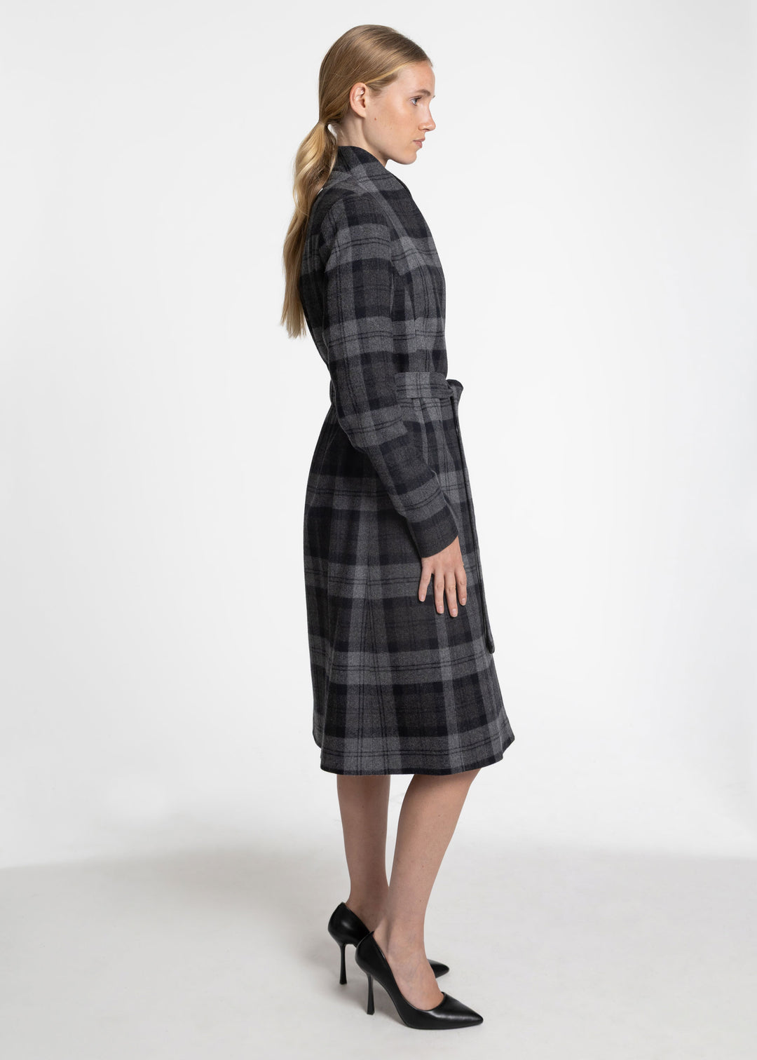 A woman modeling a long-sleeved, knee-length plaid coat in dark gray and black, shown from a side profile against a plain white studio background.