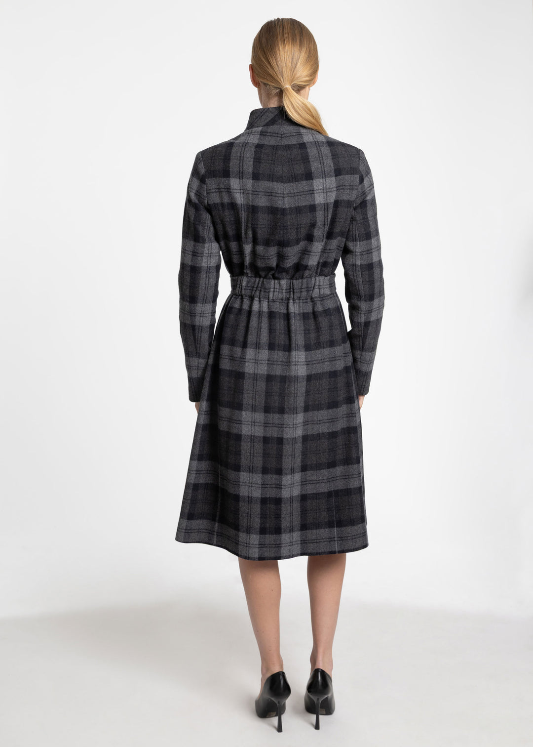 A woman viewed from the back wearing a dark gray and black plaid mid-length coat with a tie waist, paired with black heels, against a plain white studio background.