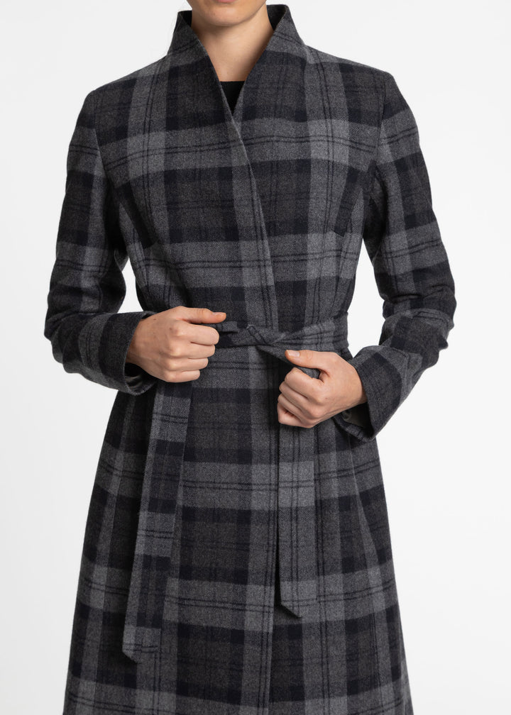 A person wearing a long plaid wrap coat in dark gray and black, with the hands holding the waist tie, against a plain light gray background.