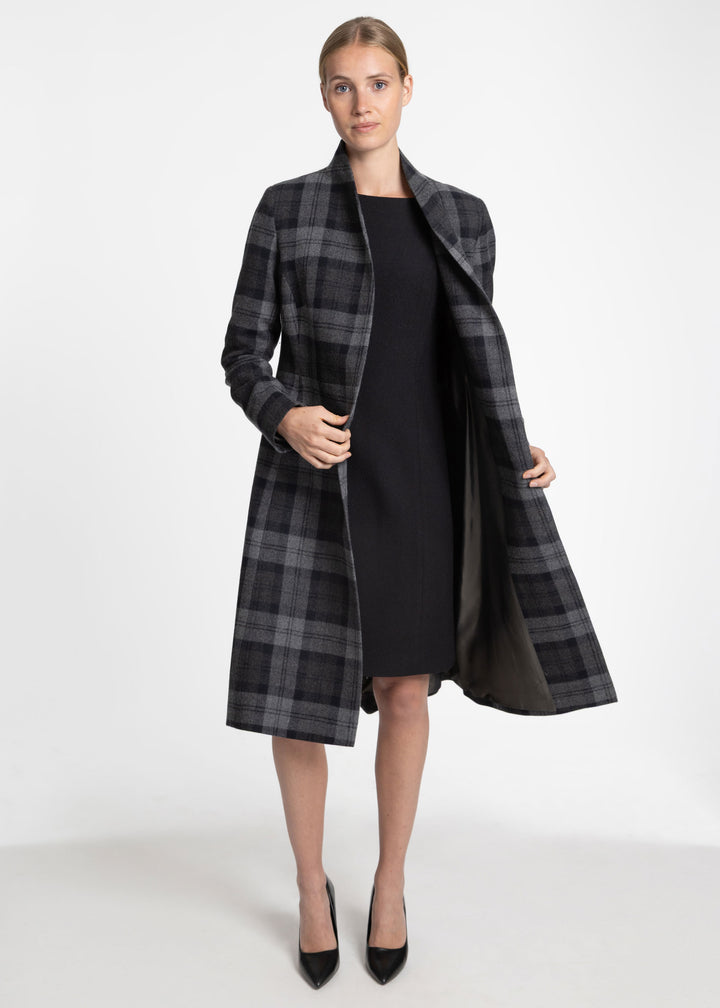 A woman models a long black and gray plaid coat over a black dress, standing against a plain white studio background.