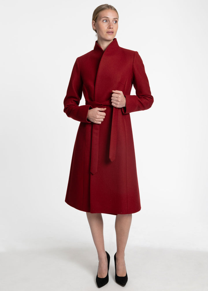 A woman modeling a long, red wrap-style coat with a high stand collar and matching tie belt, paired with black pointed-toe heels, against a plain white studio background.