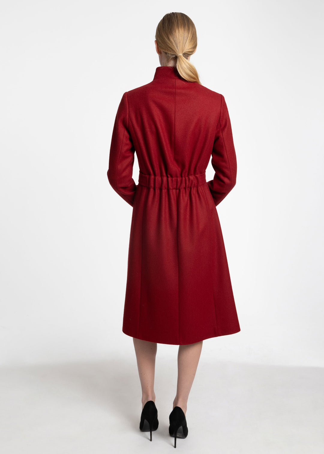 A woman viewed from the back wearing a mid-length red coat with a high stand collar, long sleeves, and a gathered waist, paired with black heels against a plain light gray studio background.
