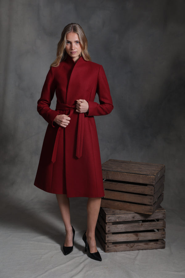 A woman models a long-sleeved, knee-length red wrap coat with a matching waist tie, standing next to a stack of rustic wooden crates against a dark gray studio backdrop.