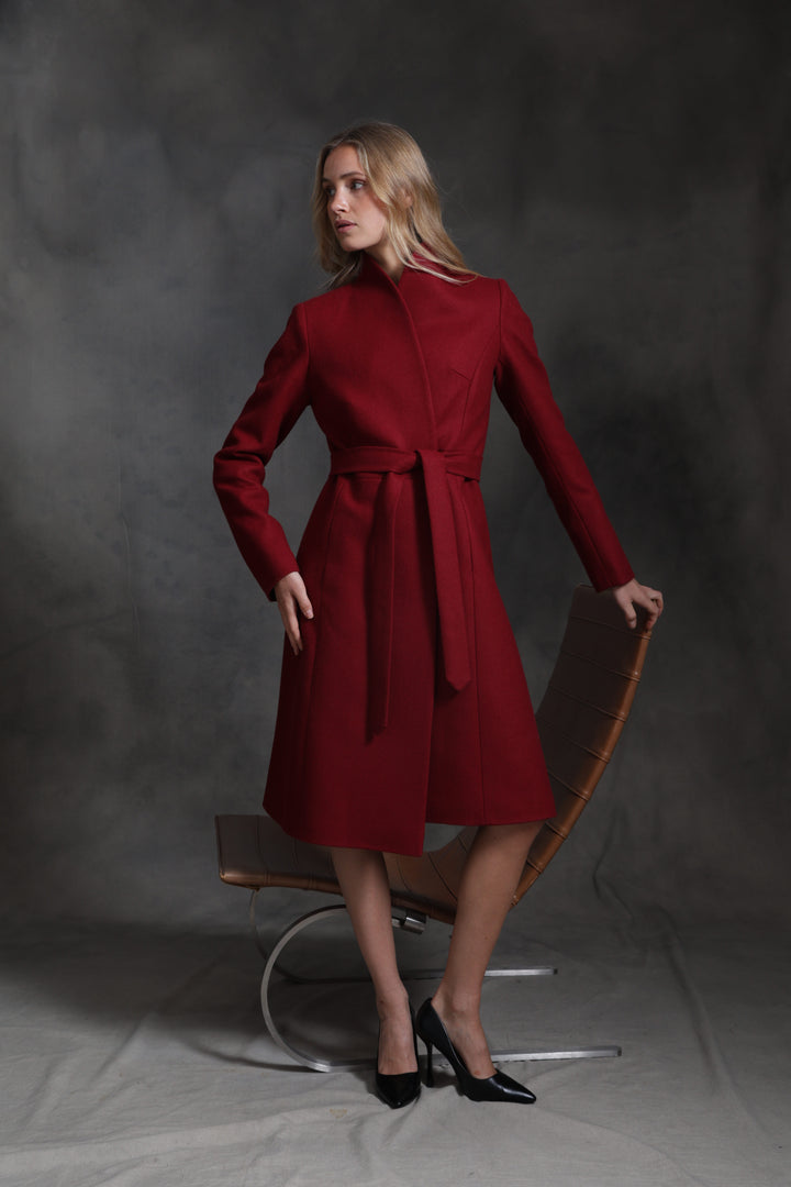 A woman models a long-sleeved, knee-length red wrap coat with a matching waist tie, standing next to a modern curved leather chair against a dark gray studio backdrop.