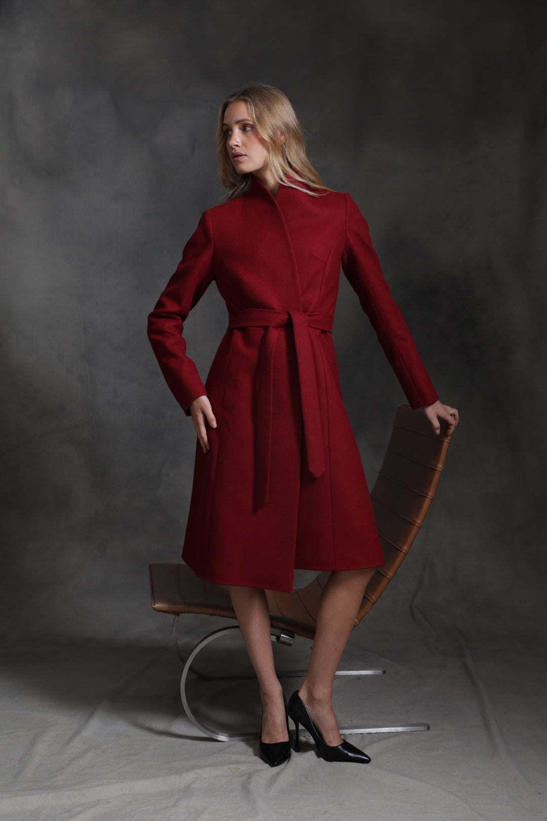 A woman models a long-sleeved, knee-length red wrap coat with a matching waist tie, standing next to a modern curved leather chair against a dark gray studio backdrop.