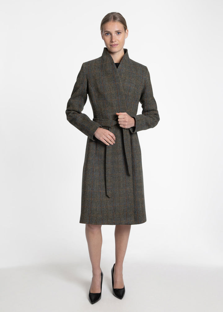A woman modeling a mid-length wrap-style coat with a shawl collar and self-tie belt, shown against a plain light gray studio background. The coat has a subtle plaid pattern in dark olive, red, and muted blue tones, and she is wearing black pointed heels.